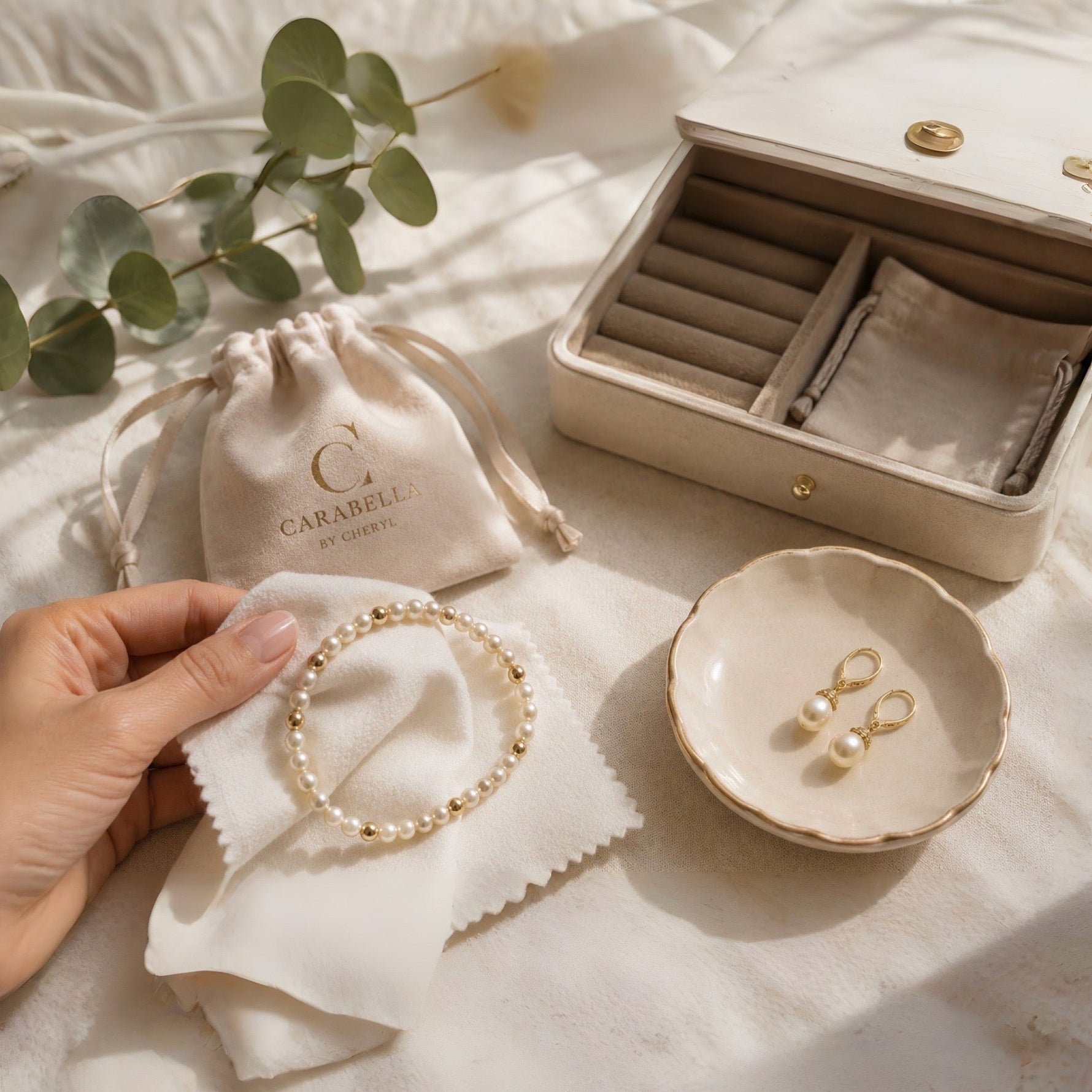 Hand gently cleaning a Carabella Swarovski® Crystal Pearl bracelet with a soft polishing cloth beside pearl earrings and a jewellery box in natural window light. 