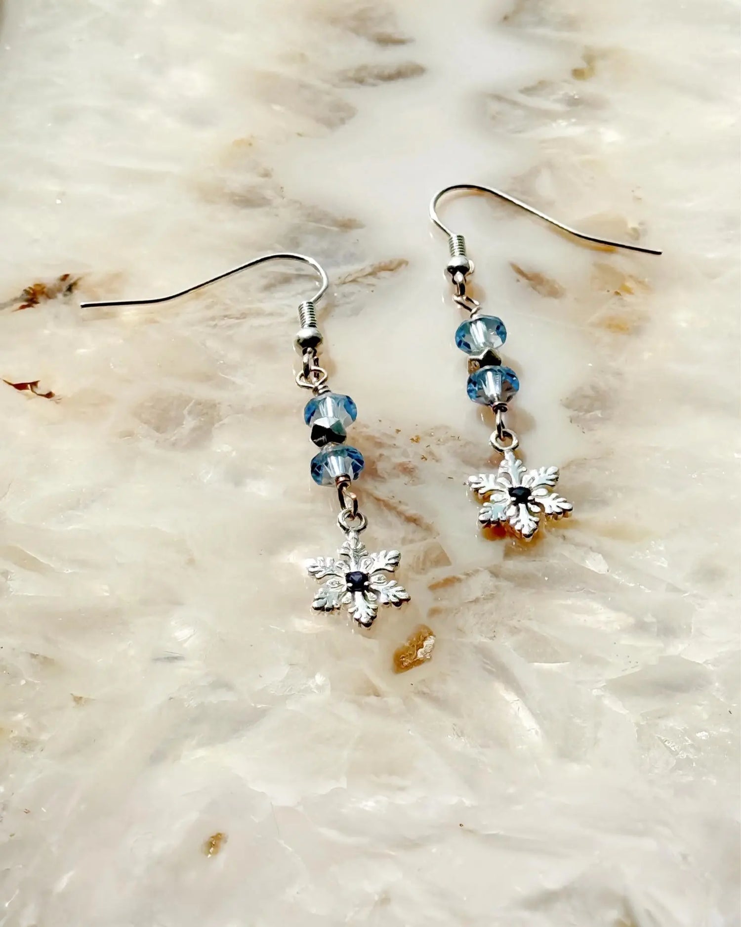 Sapphire Snowflake Earrings, handcrafted in Canada, featuring sterling silver snowflake charms, light blue European crystals, and a lab-grown sapphire centre, photographed on a luminous stone background.