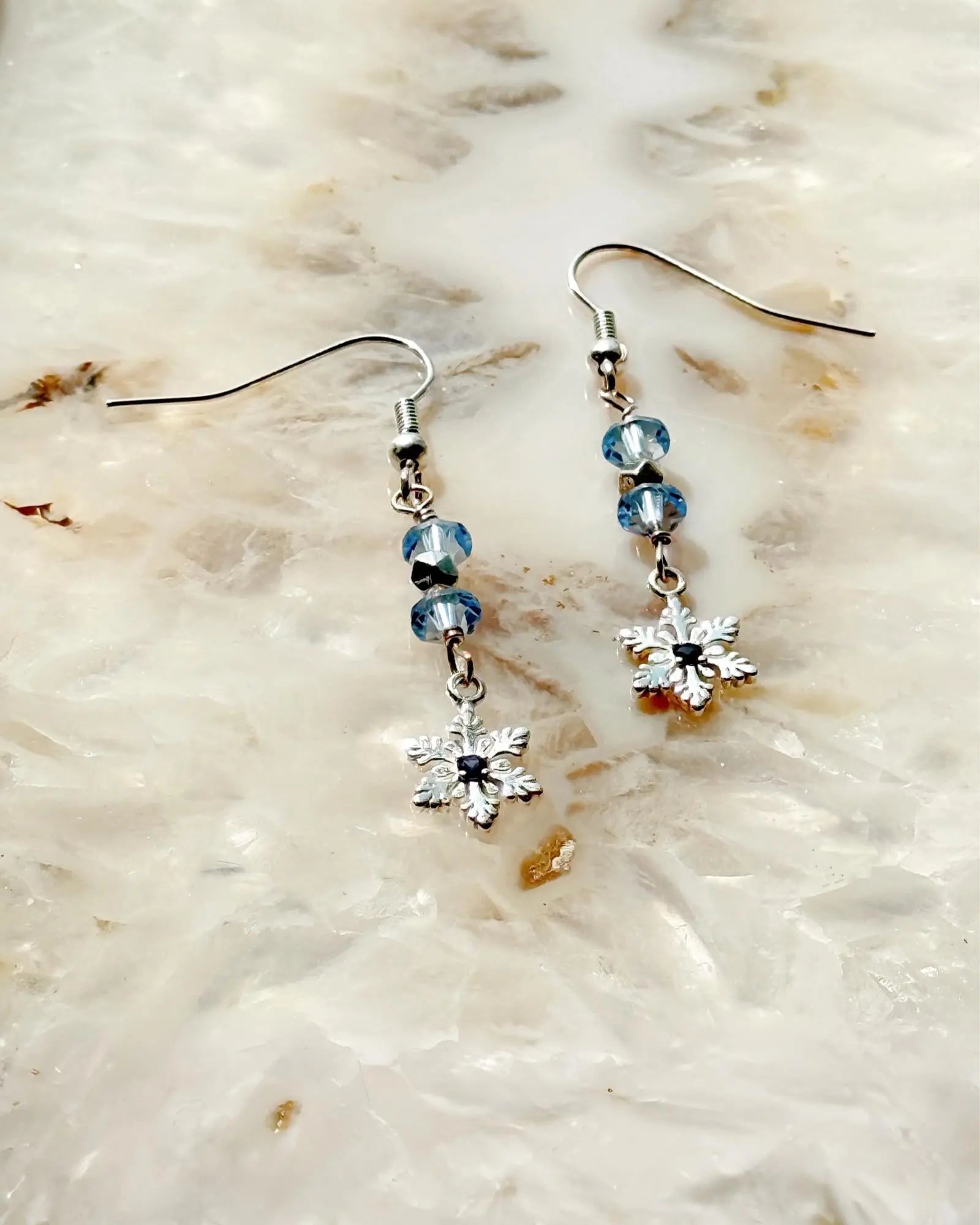 Sapphire Snowflake Earrings, handcrafted in Canada, featuring sterling silver snowflake charms, light blue European crystals, and a lab-grown sapphire centre, photographed on a luminous stone background.