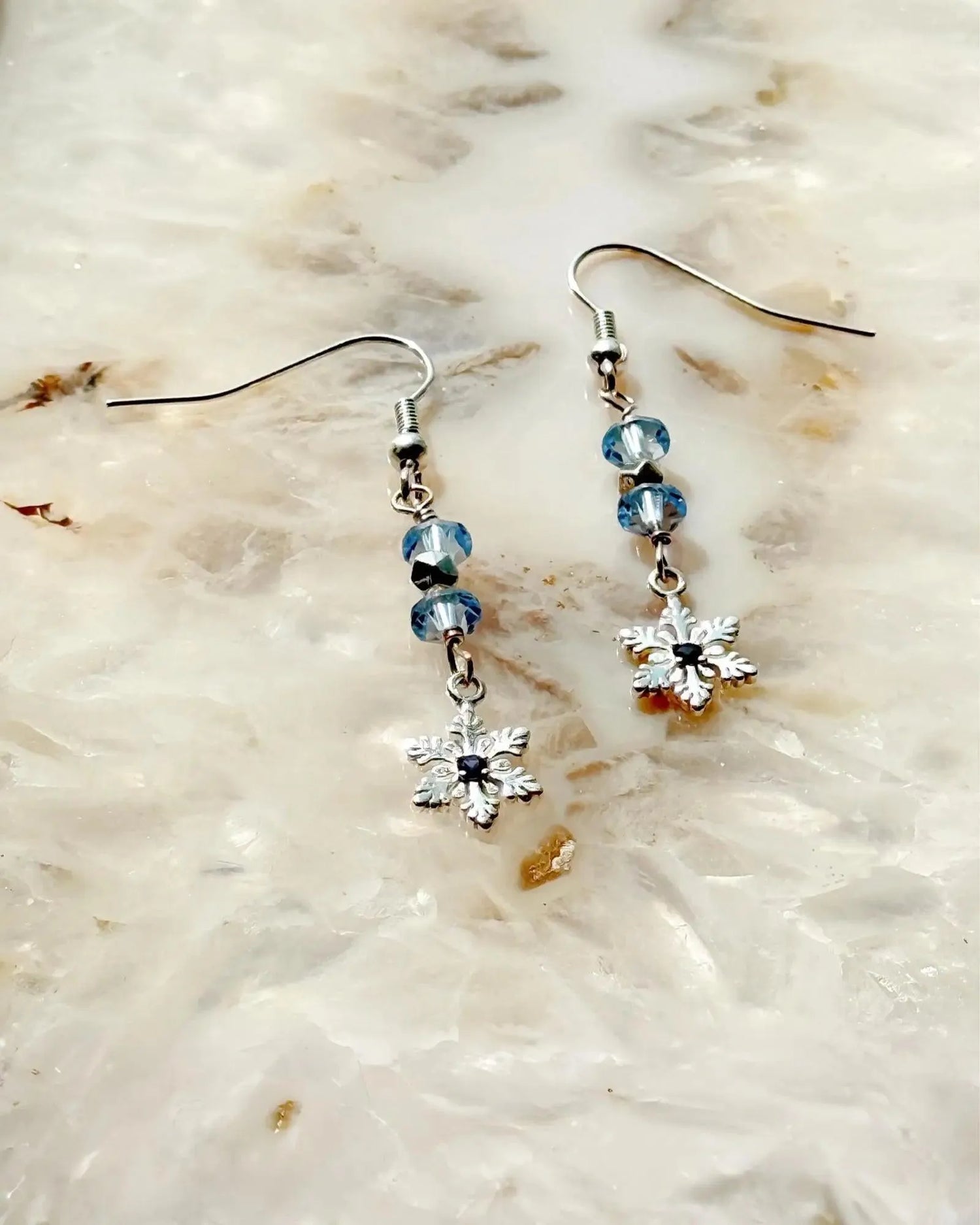 Sapphire Snowflake Earrings, handcrafted in Canada, featuring sterling silver snowflake charms, light blue European crystals, and a lab-grown sapphire centre, photographed on a luminous stone background.