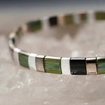 Close-up macro photograph of the Ainsley Miyuki Tila Bracelet showcasing premium Japanese Miyuki glass tila beads in green, black, white, and pewter tones arranged on white quartz crystal.