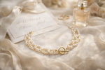 Freshwater pearl necklace with gold toggle clasp styled on ivory silk beside wedding invitation, perfume bottle and rings in an elegant bridal editorial flat lay.