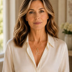 Woman wearing the Cara gold Heart B necklace with an ivory silk blouse in a refined luxury interior.
