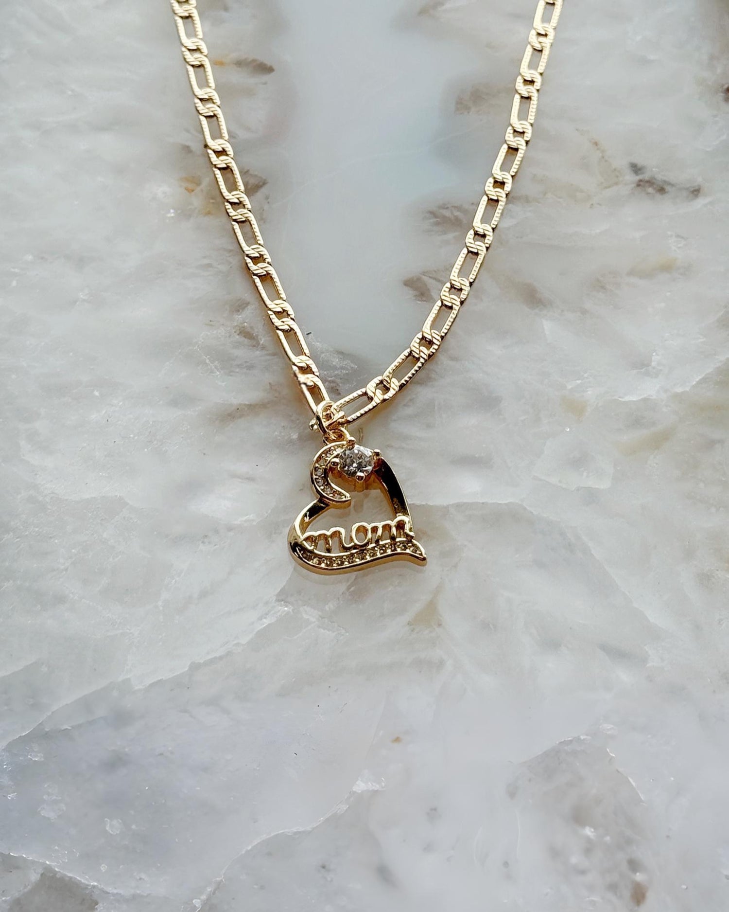 Eternal Love “Mom” heart necklace in gold-plated finish with cubic zirconia accents, styled on soft stone for a timeless and elegant Carabella by Cheryl presentation.