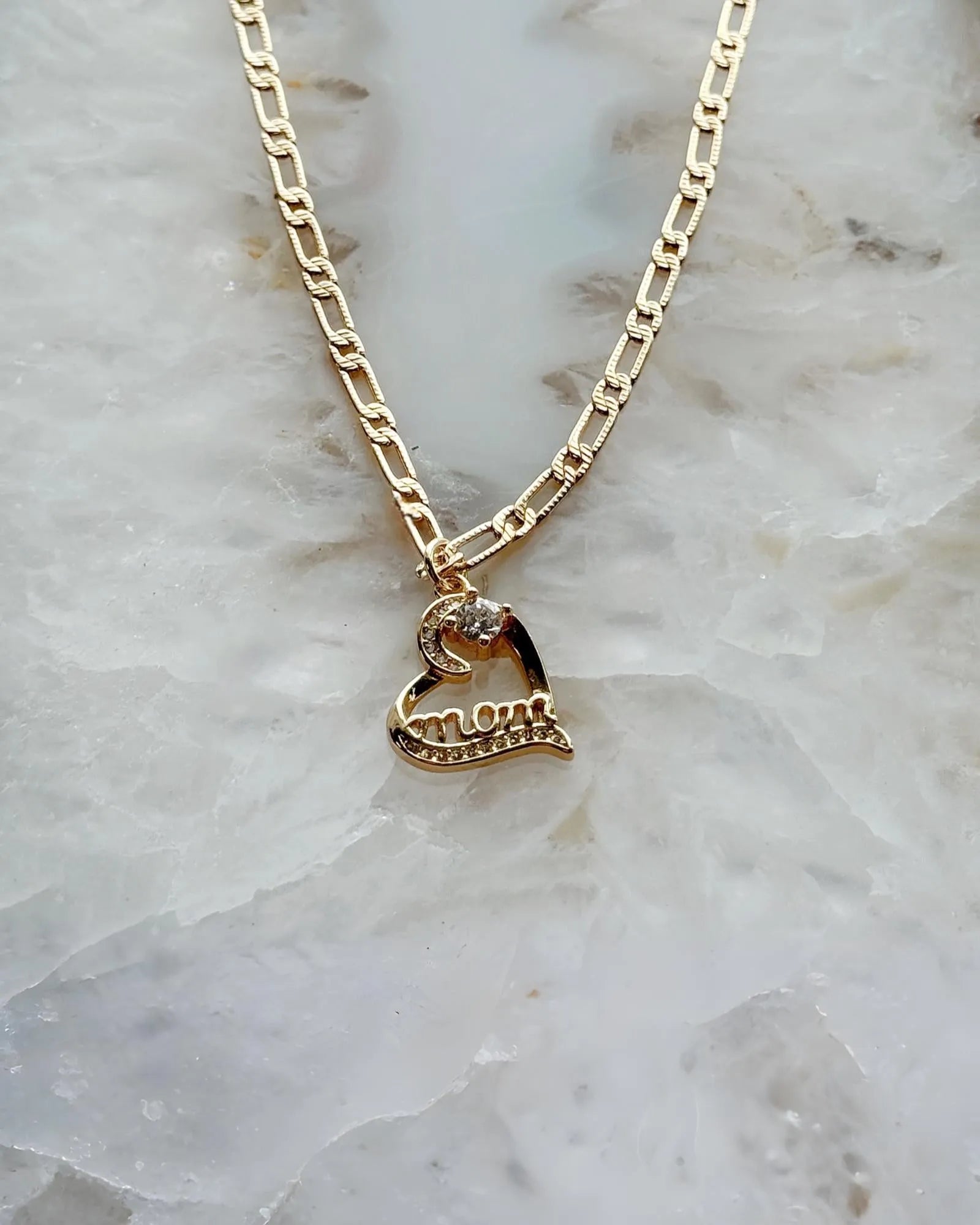 Eternal Love “Mom” heart necklace in gold-plated finish with cubic zirconia accents, styled on soft stone for a timeless and elegant Carabella by Cheryl presentation.