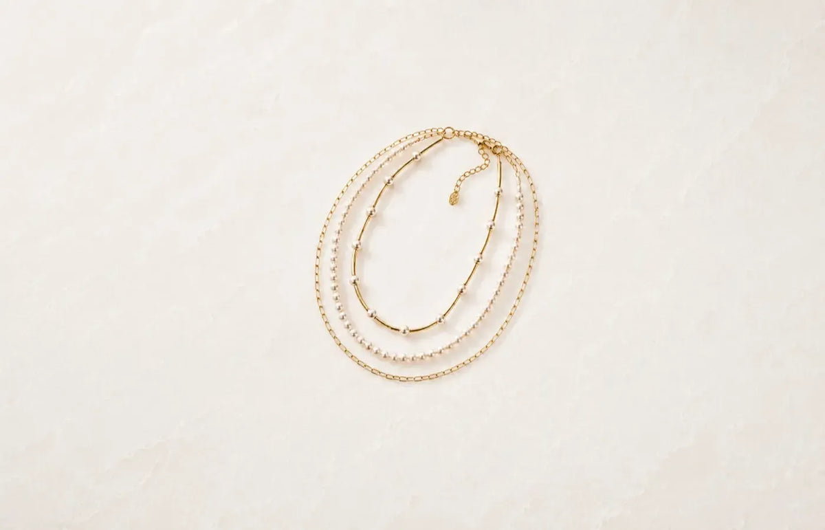 Layered gold and pearl necklaces arranged on a soft white stone background for the Carabella necklace collection.