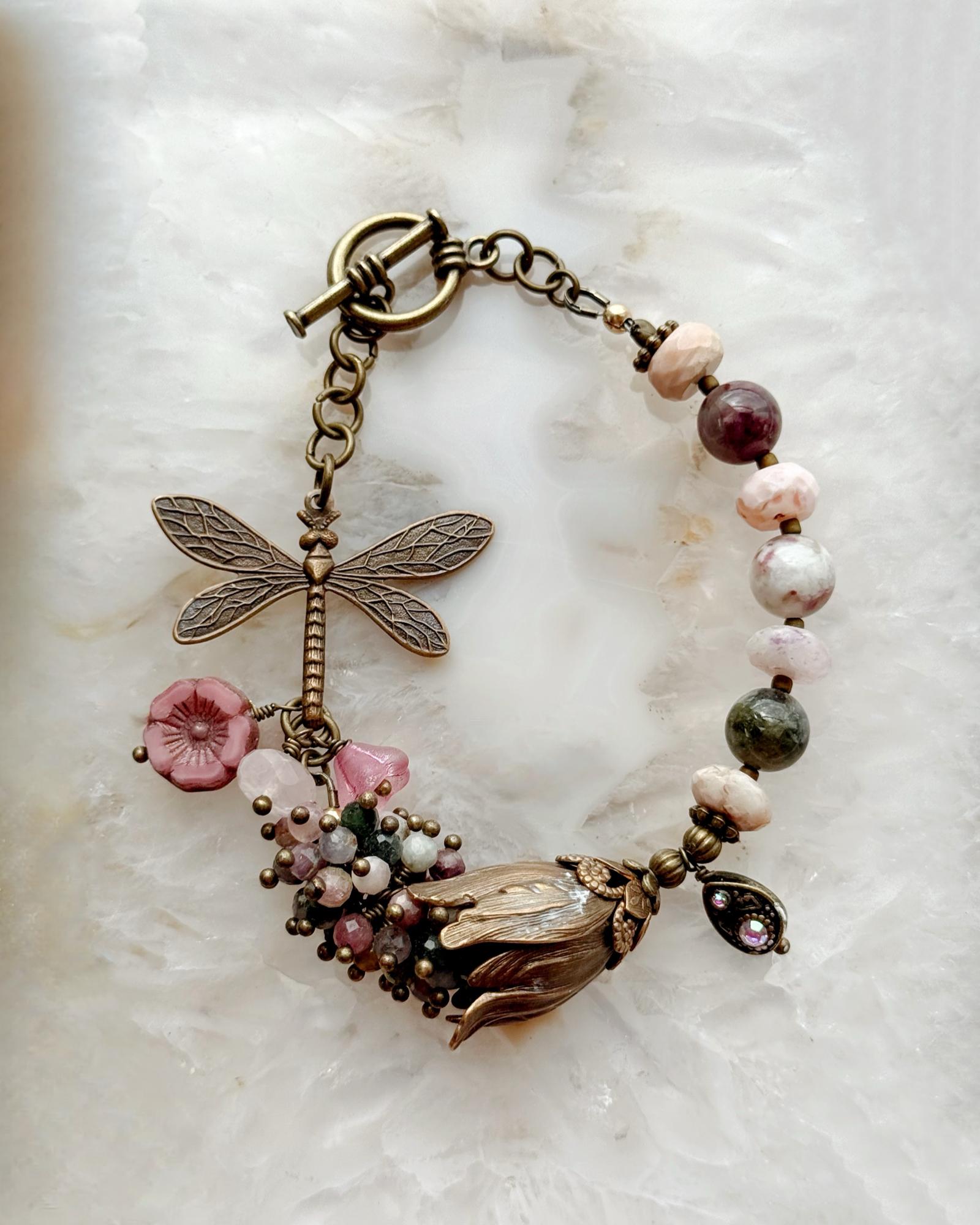 Dragonfly Garden Reverie Bracelet by Carabella by Cheryl, featuring Vintaj brass, pink tourmaline gemstones, floral accents, and a botanical charm cluster displayed on a white crystal background