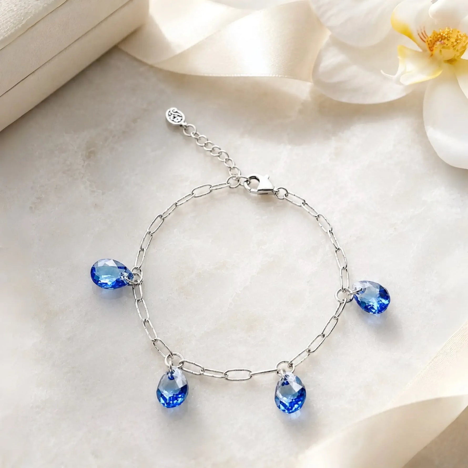 Ella silver charm bracelet with sapphire blue crystal drops presented on white quartz with ivory ribbon and white orchid in an elegant luxury still life.