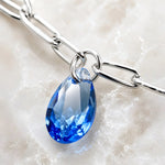 Macro detail of the Ella bracelet showing a sapphire blue crystal pear charm, polished silver paperclip chain, and handcrafted construction.