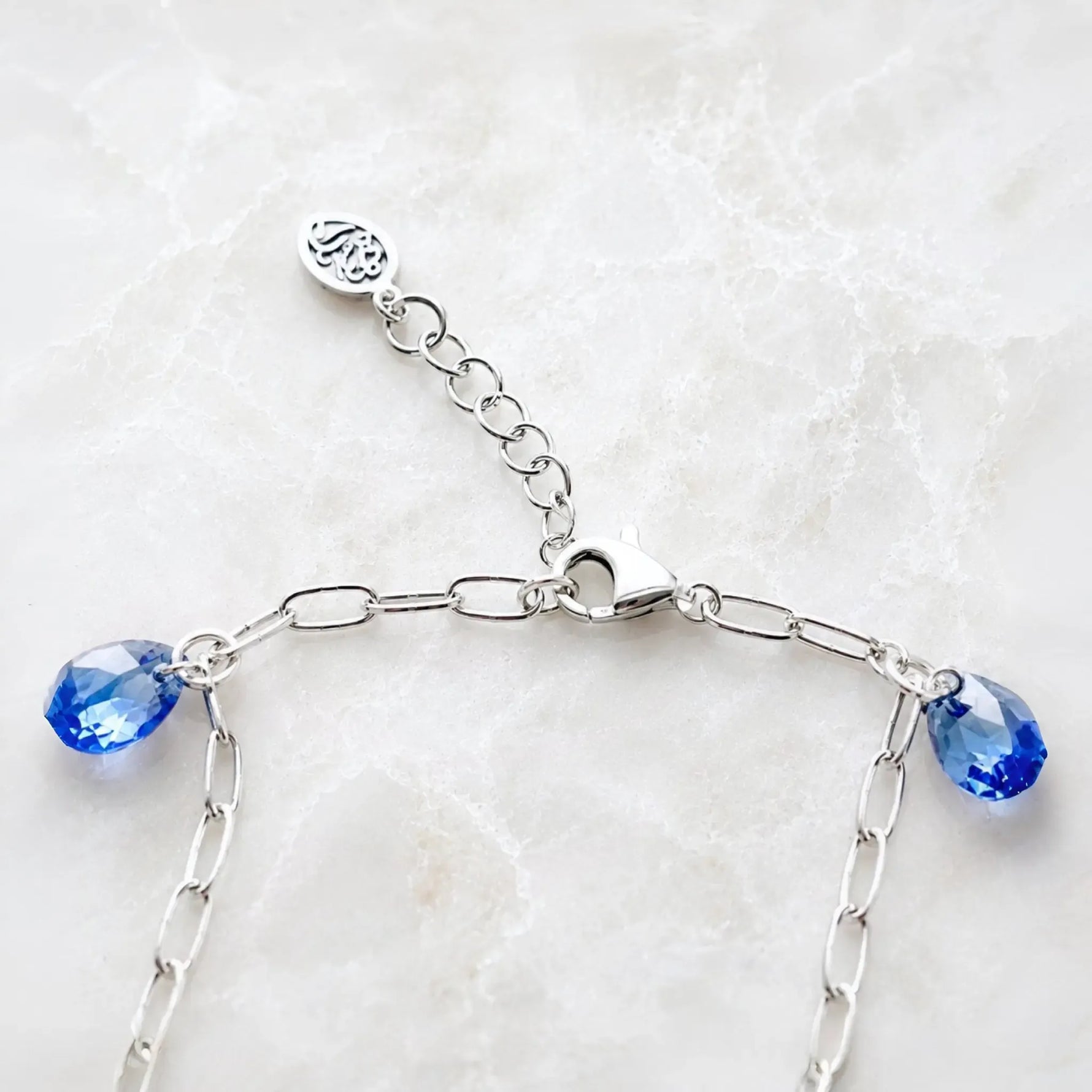 Rear clasp detail of the Ella silver charm bracelet featuring the polished lobster clasp, adjustable extender chain, signature tag, and sapphire blue crystal drops handcrafted in sterling silver.