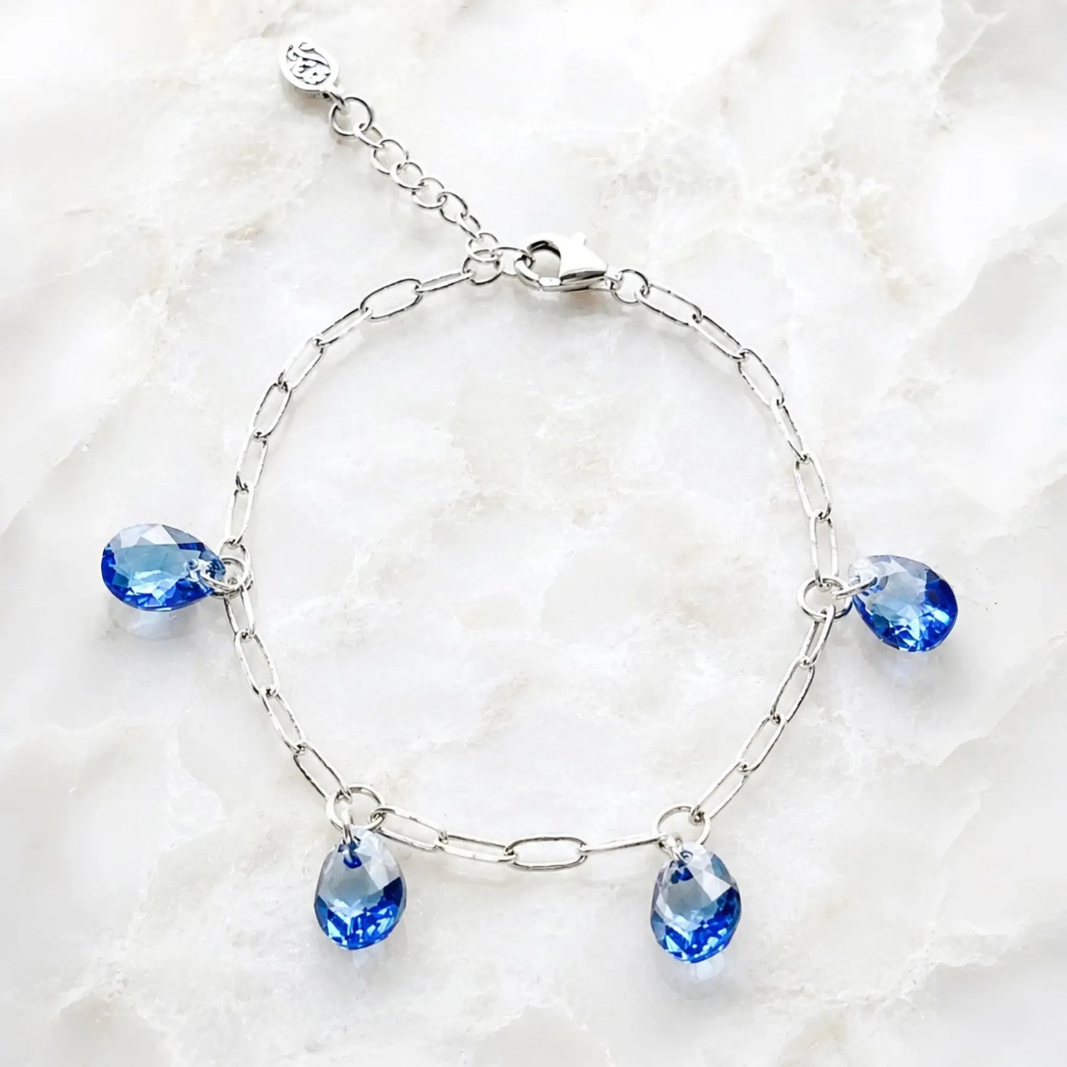 Ella silver charm bracelet with four sapphire blue crystal pear drops displayed on white quartz.