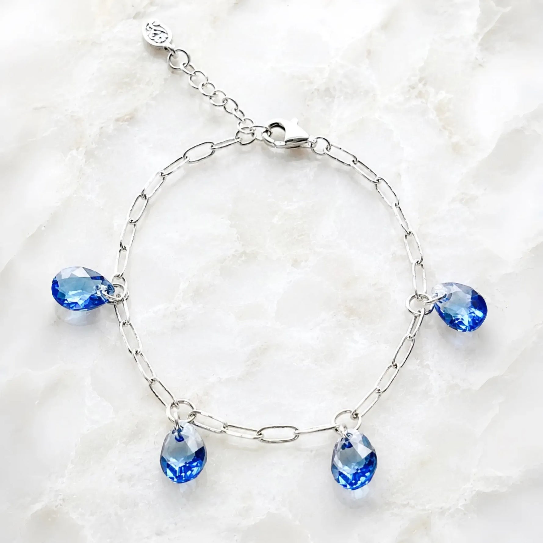 Ella silver charm bracelet with four sapphire blue crystal pear drops displayed on white quartz.