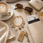 Esme Gold Charm Bracelet styled with a passport, sunglasses, cappuccino, and travel essentials, highlighting handcrafted luxury and timeless travel style.