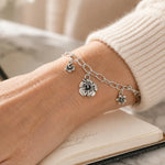 Editorial close-up of the Flora handwoven .999 fine silver bracelet with three floral pewter charms worn on the wrist of an elegant woman, paired with a cream cashmere sweater and notebook in soft natural light.