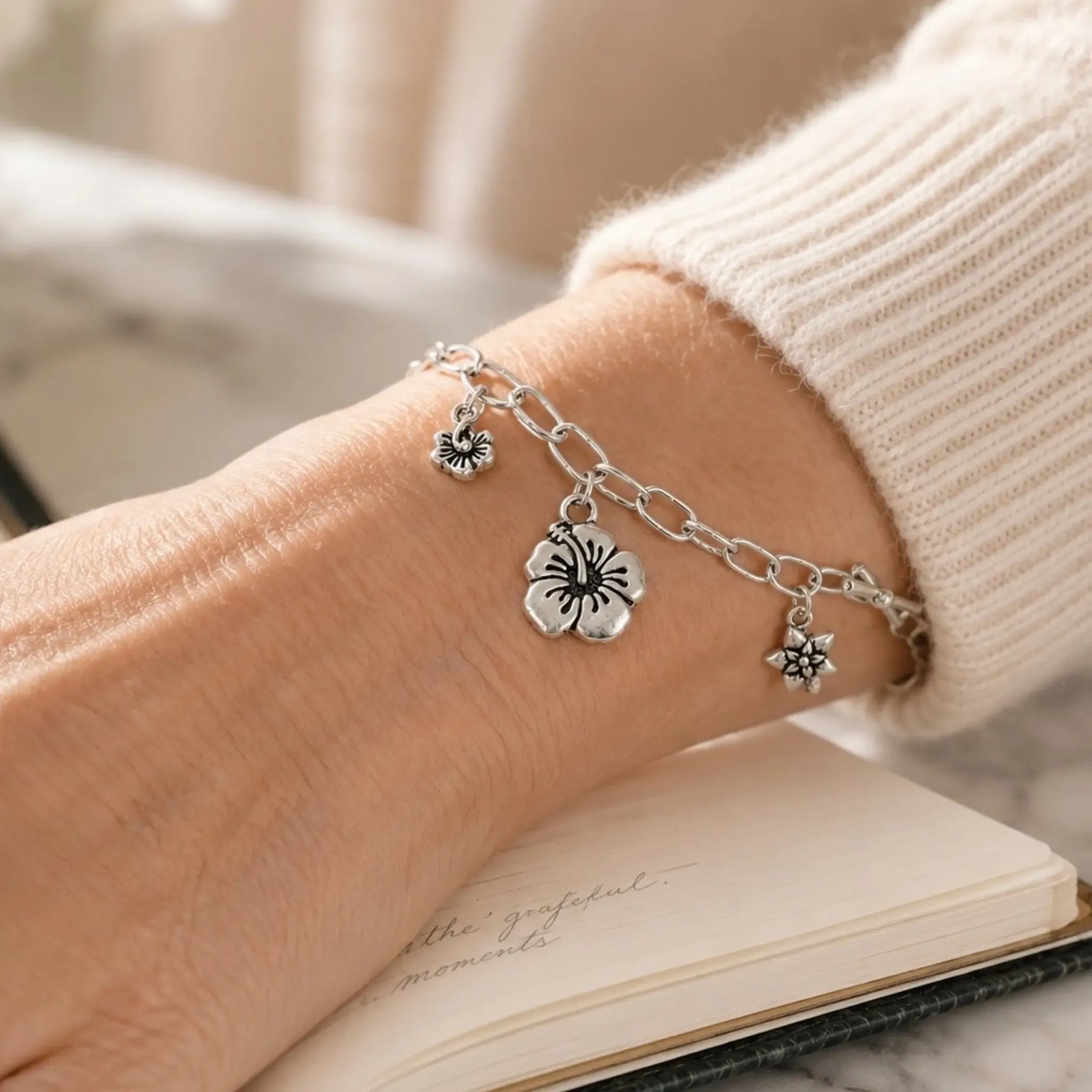 Editorial close-up of the Flora handwoven .999 fine silver bracelet with three floral pewter charms worn on the wrist of an elegant woman, paired with a cream cashmere sweater and notebook in soft natural light.