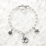Flora handwoven .999 fine silver bracelet with three floral pewter charms displayed on luminous white quartz, handcrafted in Canada.