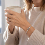Close-up of the Gemma handwoven .999 fine silver bracelet worn with a cream cashmere sweater while holding a coffee mug in soft morning light.