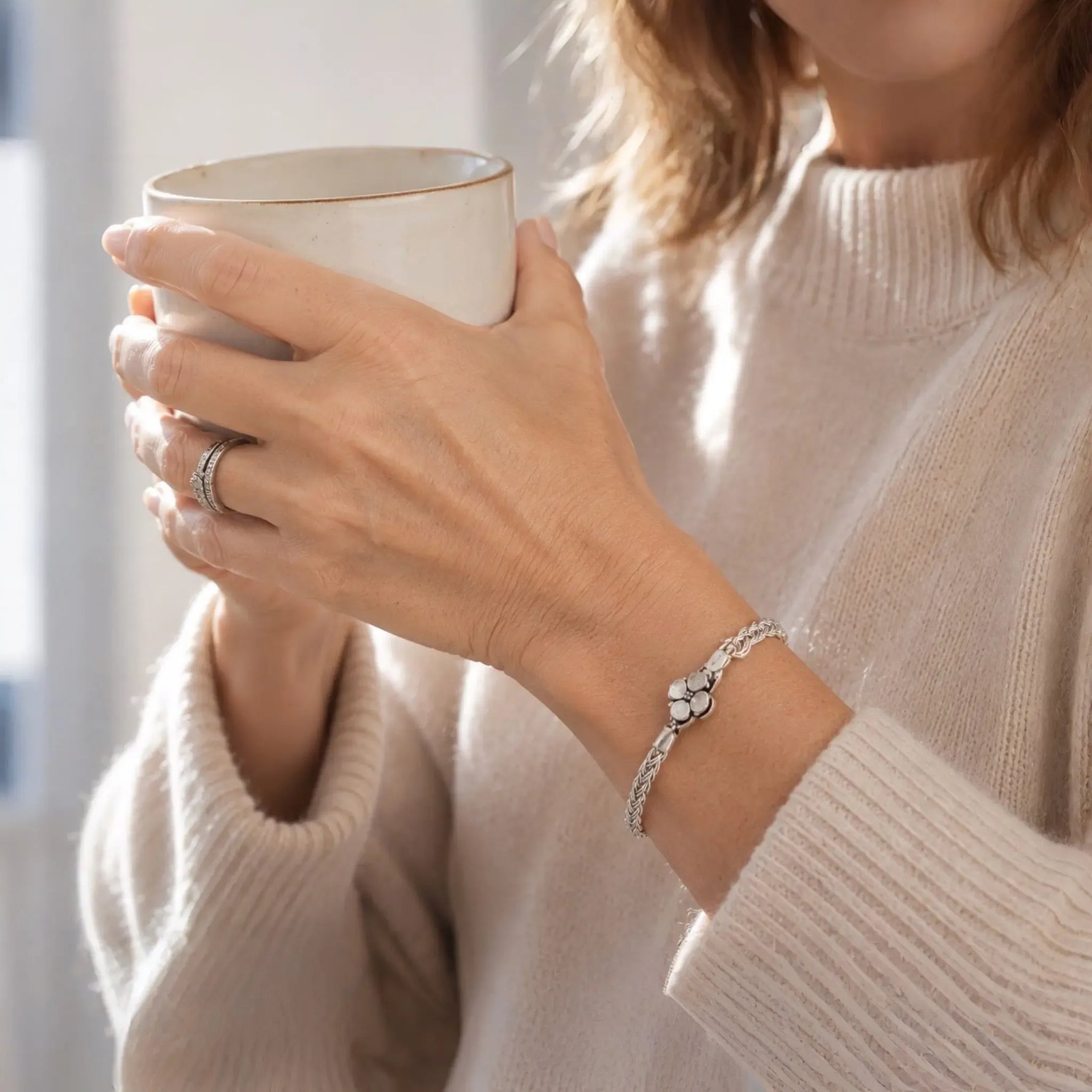 Close-up of the Gemma handwoven .999 fine silver bracelet worn with a cream cashmere sweater while holding a coffee mug in soft morning light.