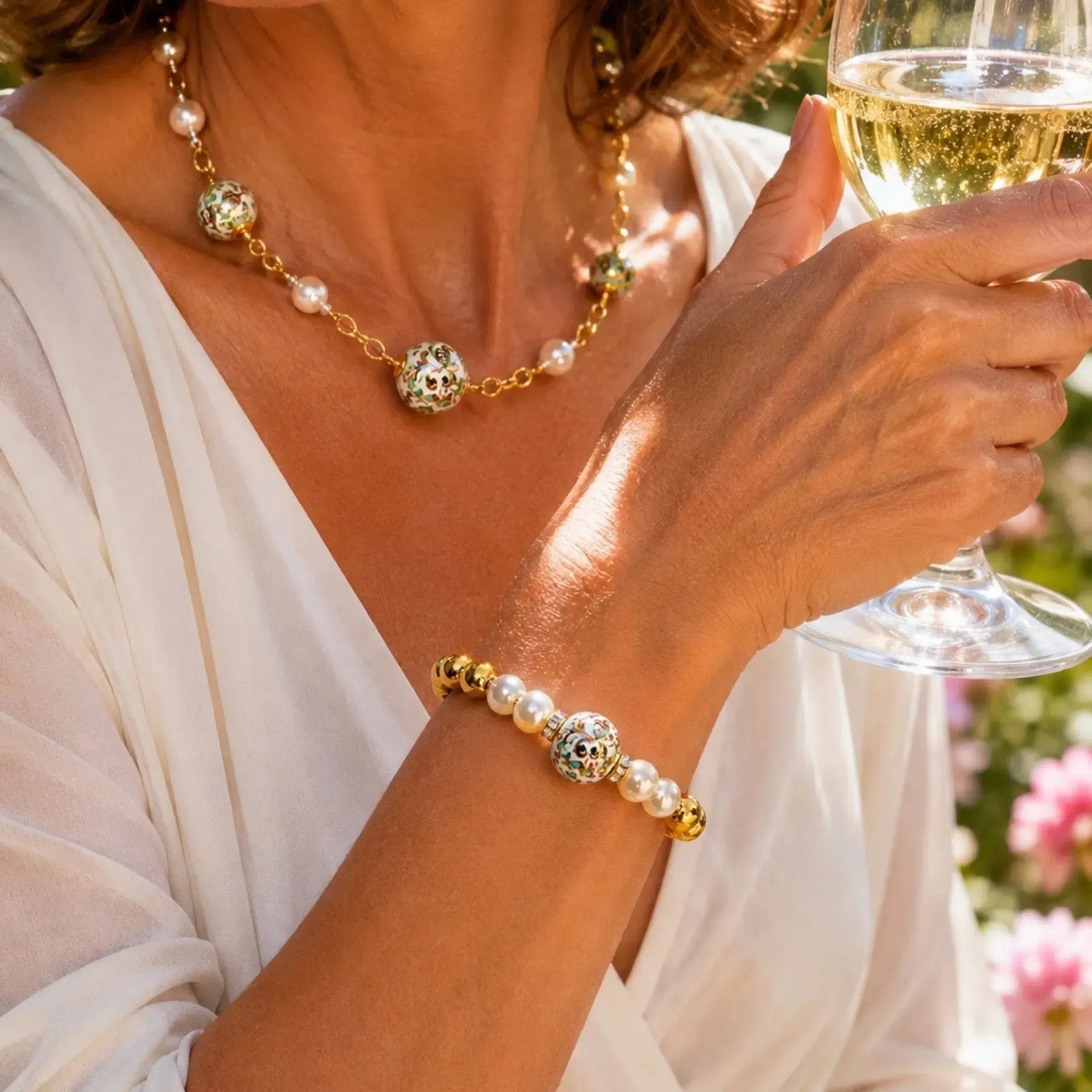 Elegant woman wearing the Imperial Blossom Necklace and Imperial Blossom Bracelet featuring rare Japanese Tensha beads, Swarovski® Crystal Pearls, and gold accents while holding a glass of wine at a summer garden party.