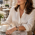 Jan and Juanita Swarovski® Crystal Pearl Bracelets layered together on the wrist of an elegant woman at a European café, showcasing coordinated black, cream, and dark grey Swarovski® Crystal Pearls with sparkling crystal rondelles.