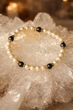 The Juanita Pearl Stretch Bracelet showcasing softly luminous pearls, charcoal accents, and gold detailing, photographed in an elevated flat lay for quiet luxury styling.