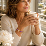 Woman wearing the Kalei amethyst bracelet while enjoying a morning cappuccino in a luxury café, showcasing violet gemstones and polished gold details.