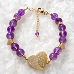 Kalei amethyst gemstone bracelet with pavé heart connector, Austrian crystal rondelles, gold heart charms, and polished gold clasp displayed on white quartz.