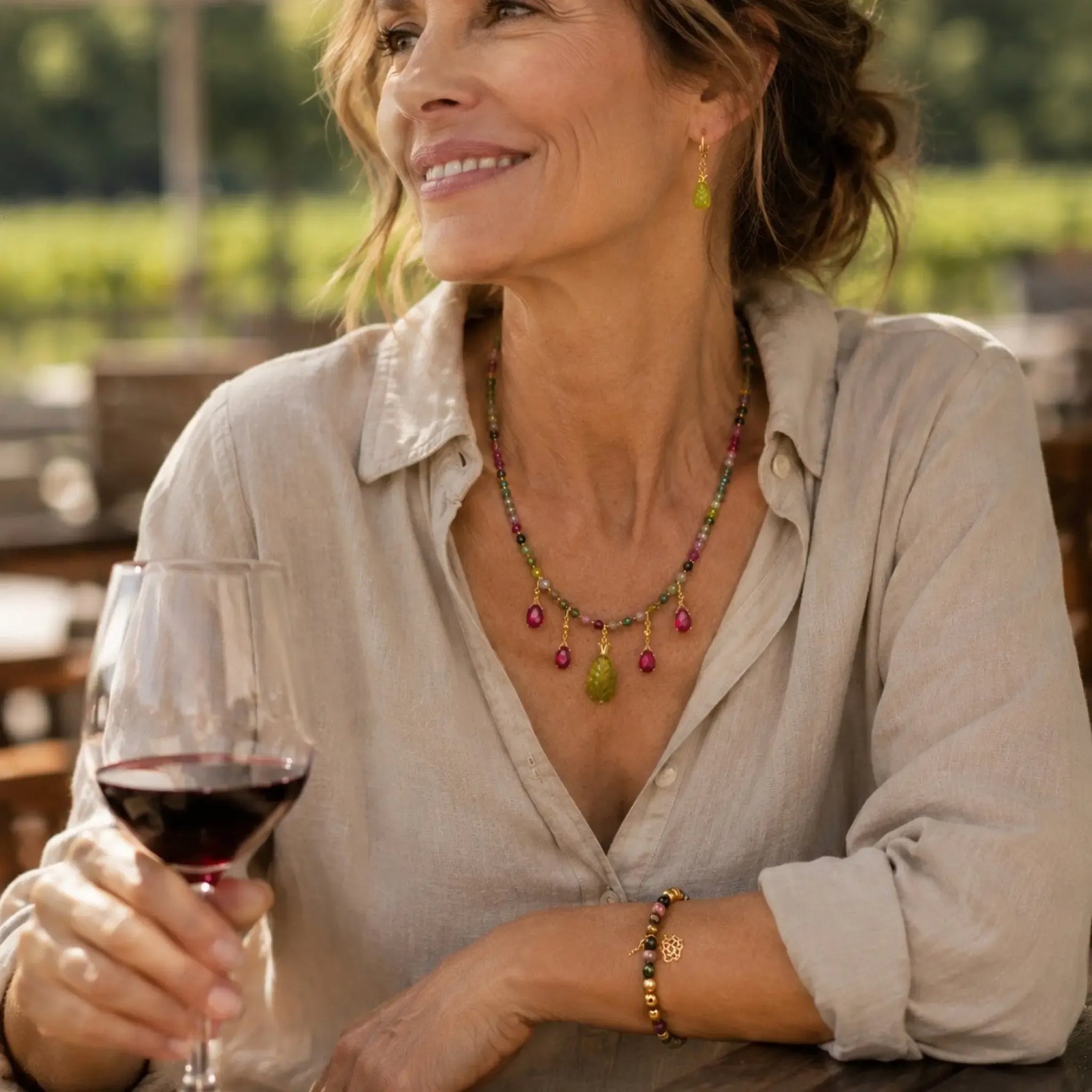 Woman wearing the Layla Rainbow Tourmaline Necklace, Layla Tourmaline Bracelet, and Layla Jade Earrings while enjoying a glass of wine at an outdoor winery.