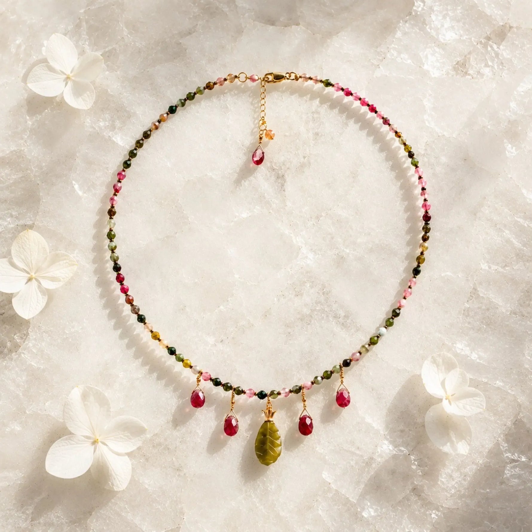 Layla Rainbow Tourmaline Necklace featuring faceted rainbow tourmaline gemstones, ruby gemstone drop charms, a carved jade pendant, gold-filled findings, and an adjustable extender chain, photographed on a quartz stone background.