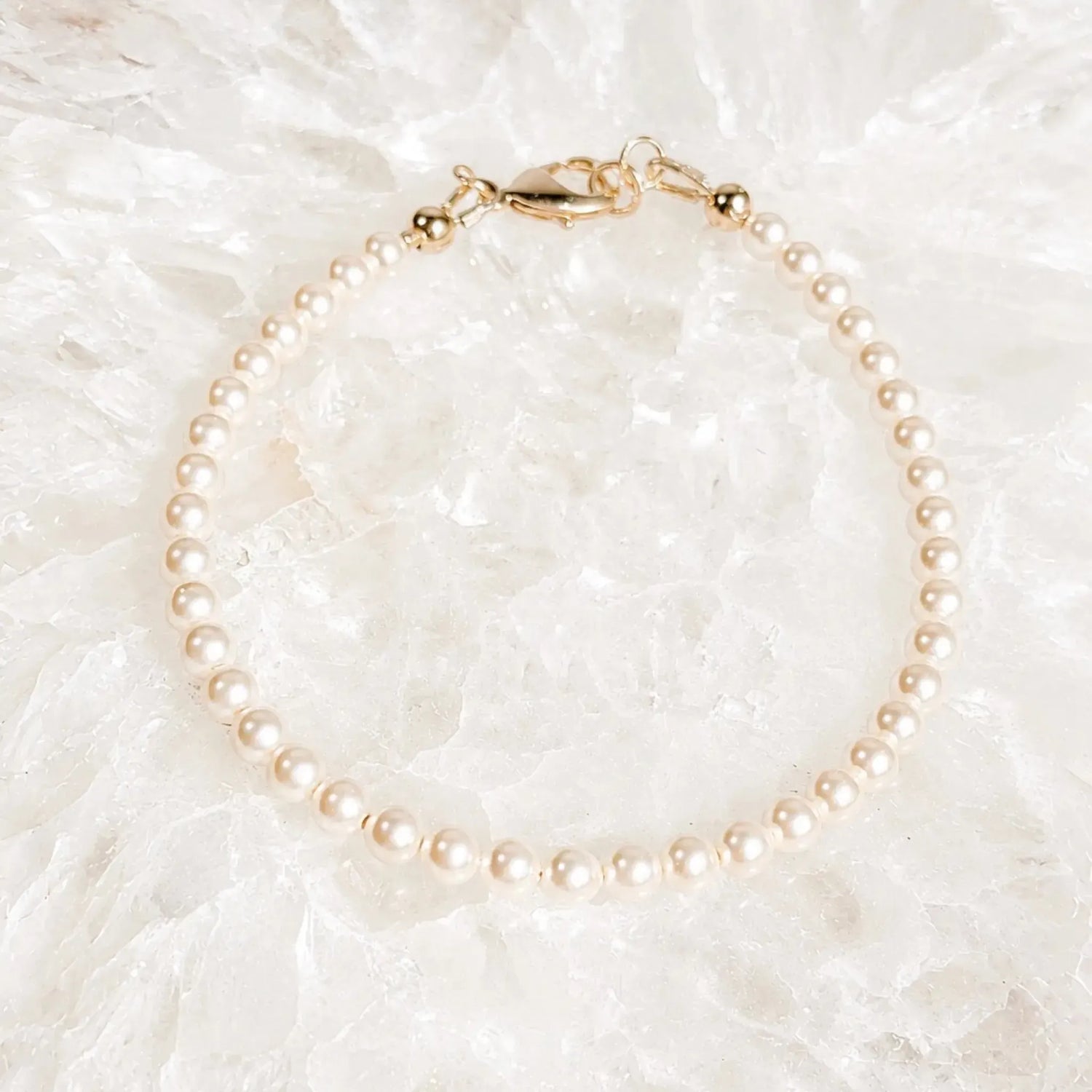 Leilani Swarovski® Crystal Pearl Bracelet resting on luminous white quartz crystal, featuring cream Swarovski® Crystal Pearls and a polished 14k gold clasp in a bright luxury flat lay.