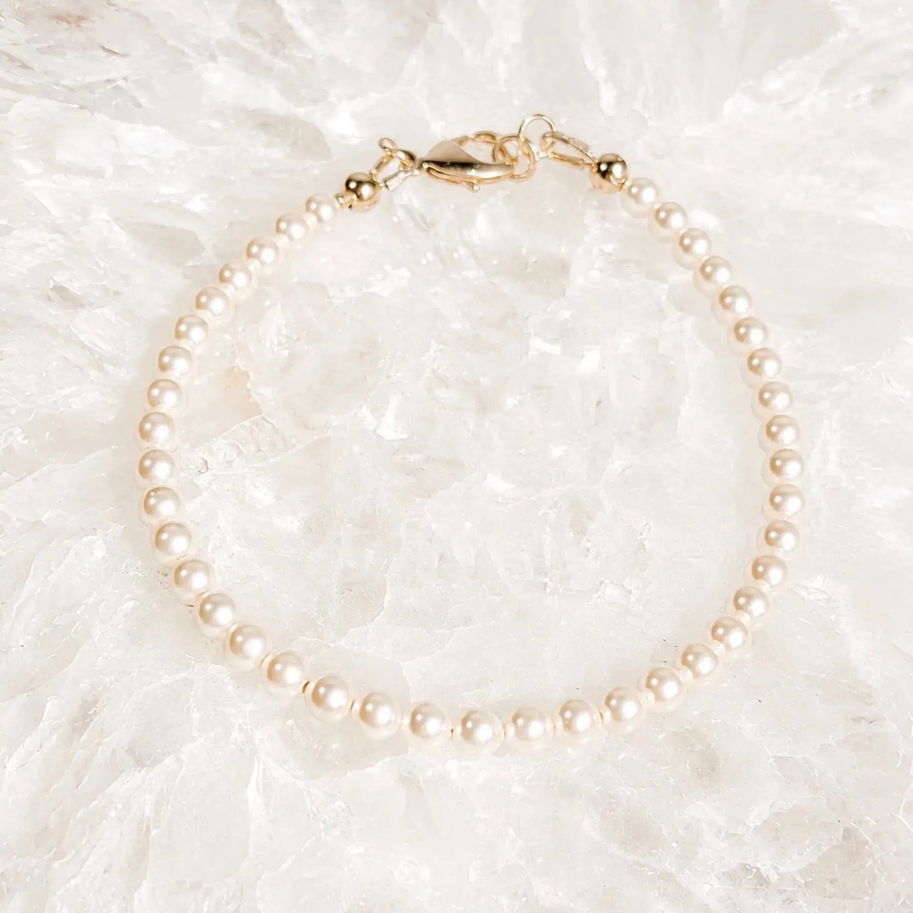 Leilani Swarovski® Crystal Pearl Bracelet resting on luminous white quartz crystal, featuring cream Swarovski® Crystal Pearls and a polished 14k gold clasp in a bright luxury flat lay.