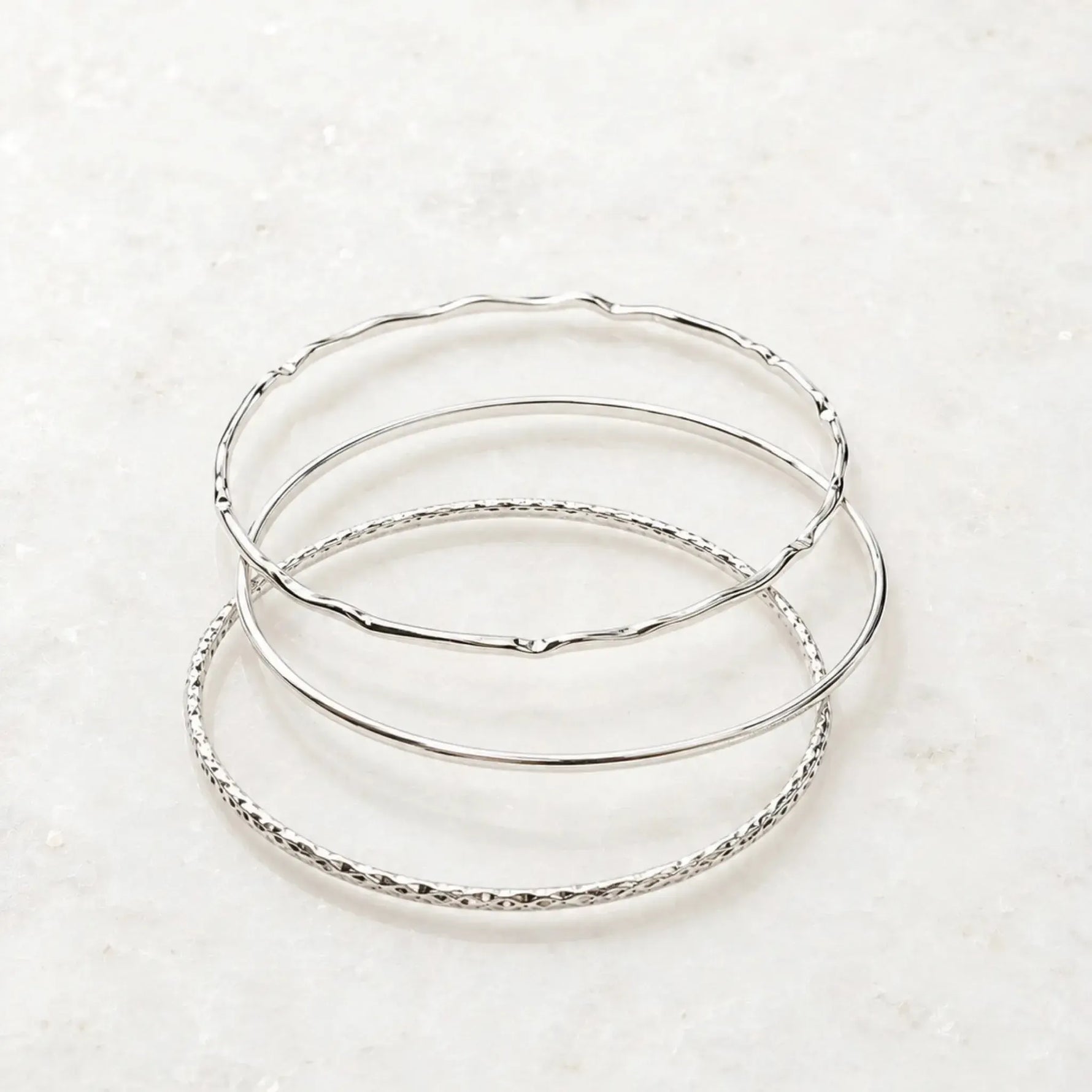 Lilith sterling silver bangles arranged on a white quartz background showcasing three handcrafted stackable bangles with polished, smooth, and hand-textured finishes.