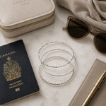 Lilith handcrafted sterling silver bangles styled with a passport, jewellery case, sunglasses, and travel accessories on a white quartz surface.