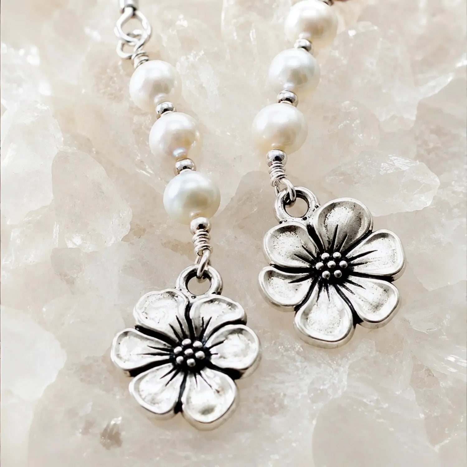 Macro detail of Mia Freshwater Pearl Earrings showcasing genuine freshwater pearls, sterling silver wire wrapping, and pewter apple blossom charms on white quartz crystal.