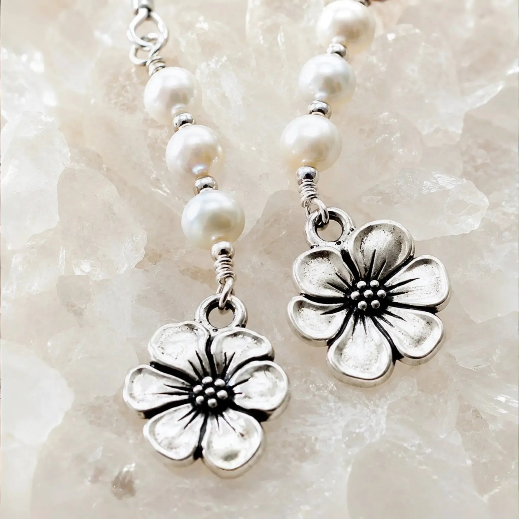 Macro detail of Mia Freshwater Pearl Earrings showcasing genuine freshwater pearls, sterling silver wire wrapping, and pewter apple blossom charms on white quartz crystal.