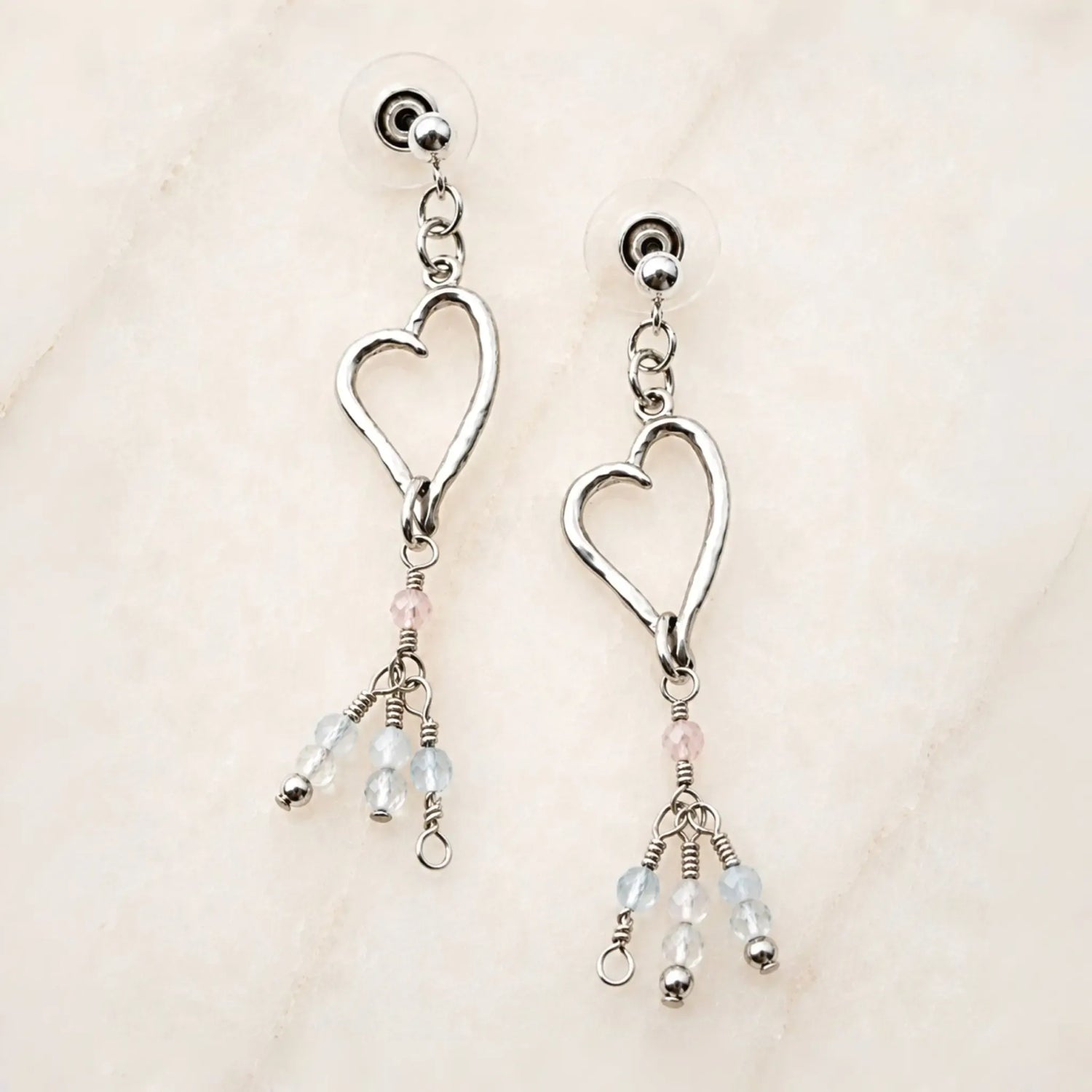 Morganite Heart Earrings handcrafted in sterling silver with blush morganite and soft blue gemstone accents on a white quartz background.