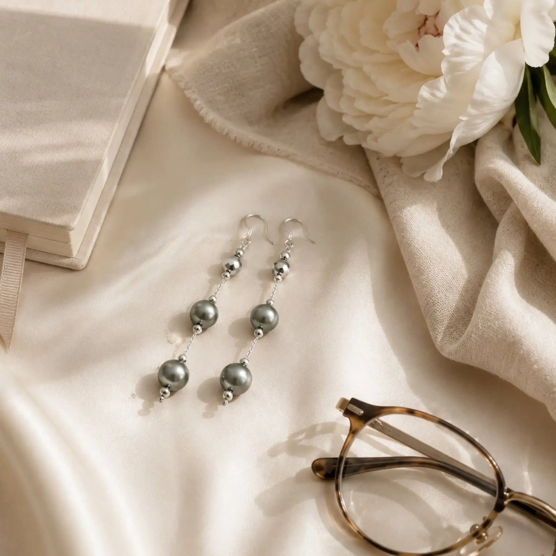Natasha Swarovski® Crystal Pearl Earrings styled on ivory silk with a cream linen scarf, white peony, vintage reading glasses, and an ivory journal in a luxury editorial flat lay.