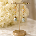 Quianna handcrafted lucky jade drop earrings displayed on an elegant gold jewellery stand with soft ivory florals, highlighting their artisan craftsmanship and luxury presentation.