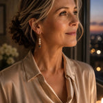 Woman wearing Serenity Swarovski® Crystal Pearl earrings with a champagne silk blouse in a refined evening setting illuminated by candlelight