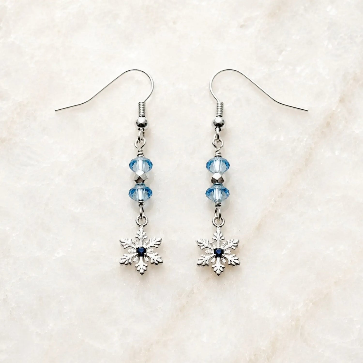Handcrafted silver snowflake earrings with sapphire-blue crystals displayed on Carabella's signature white quartz surface.