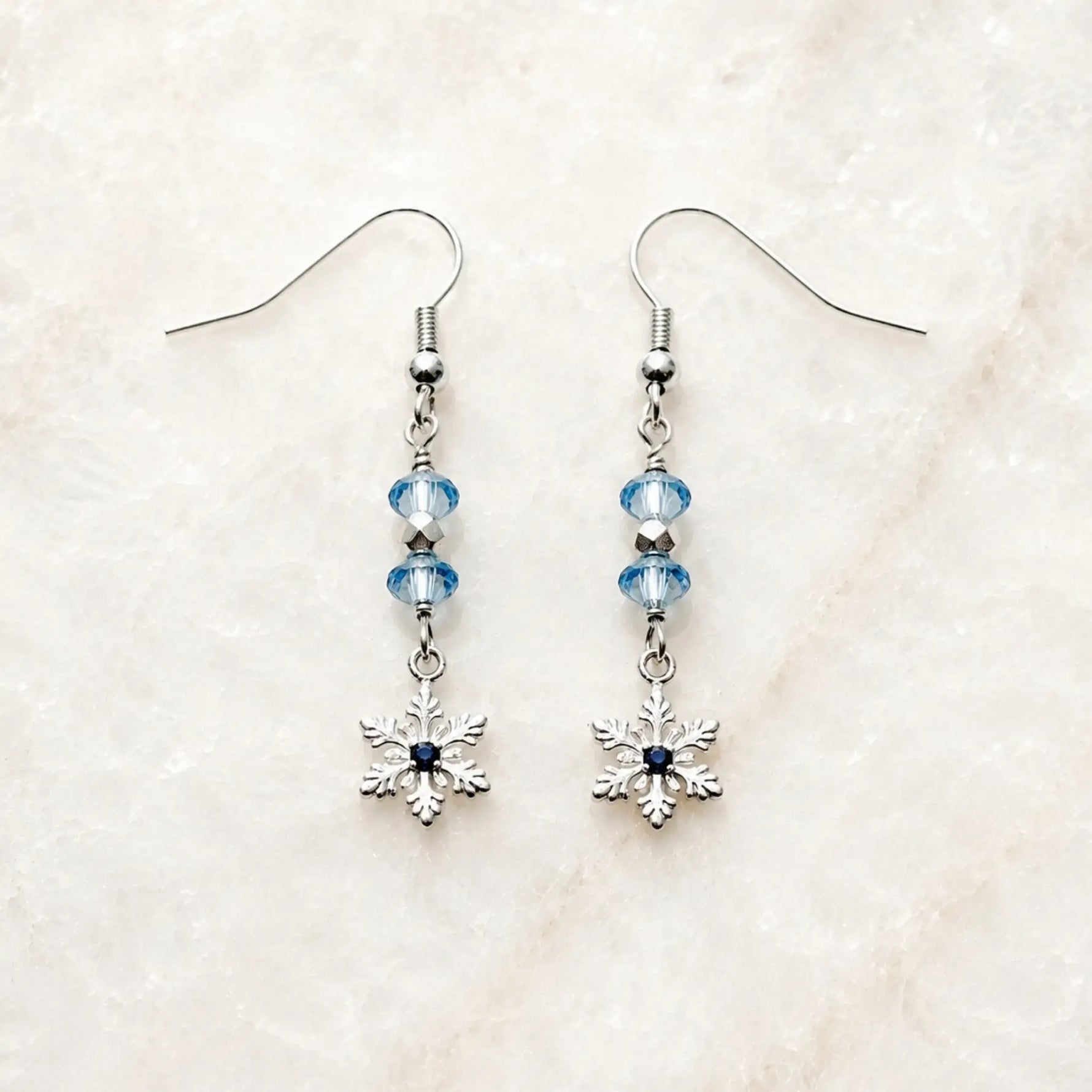 Handcrafted silver snowflake earrings with sapphire-blue crystals displayed on Carabella's signature white quartz surface.