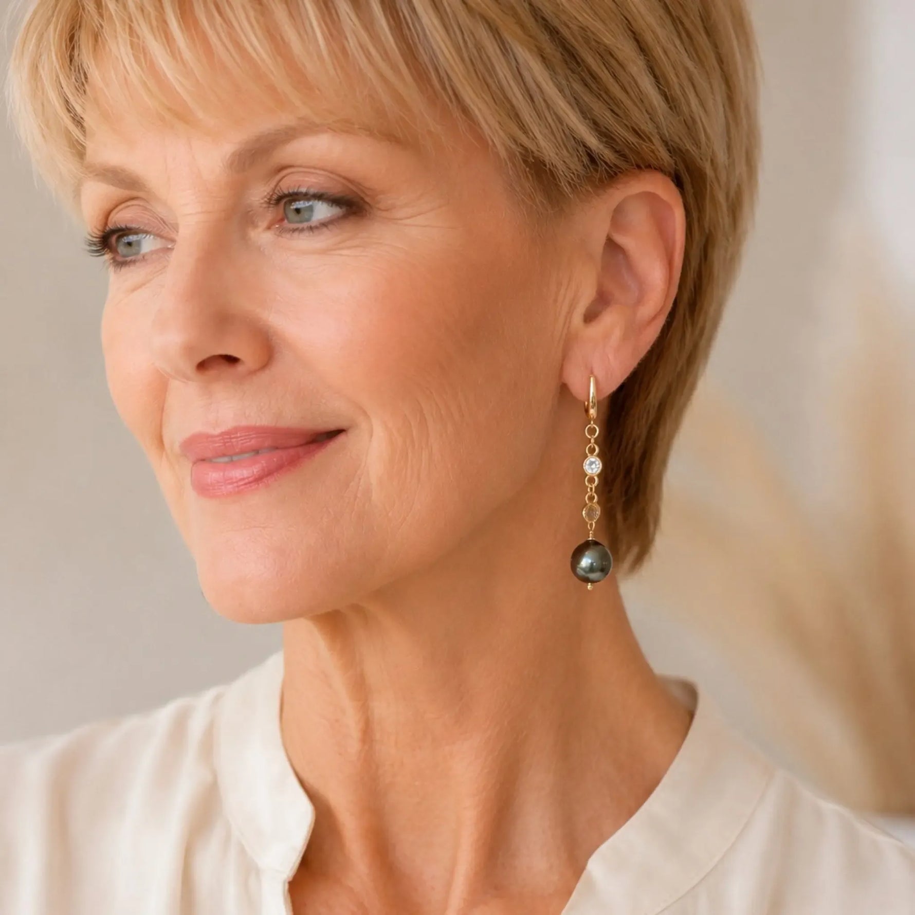 Elegant woman wearing Tahitian South Sea black pearl drop earrings with crystal accents and gold huggie hoops, styled with an ivory blouse in soft editorial lighting.