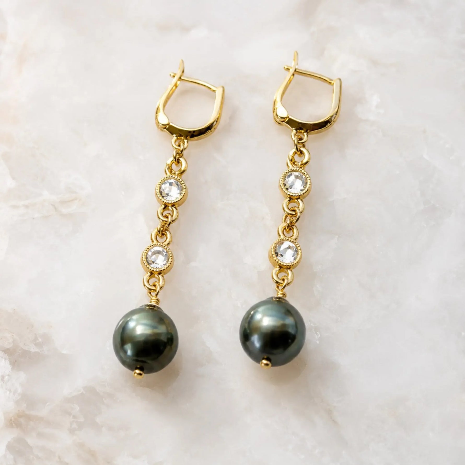 Tahitian South Sea black pearl drop earrings with crystal bezels and gold huggie hoops displayed on a white quartz background, handcrafted in Canada.
