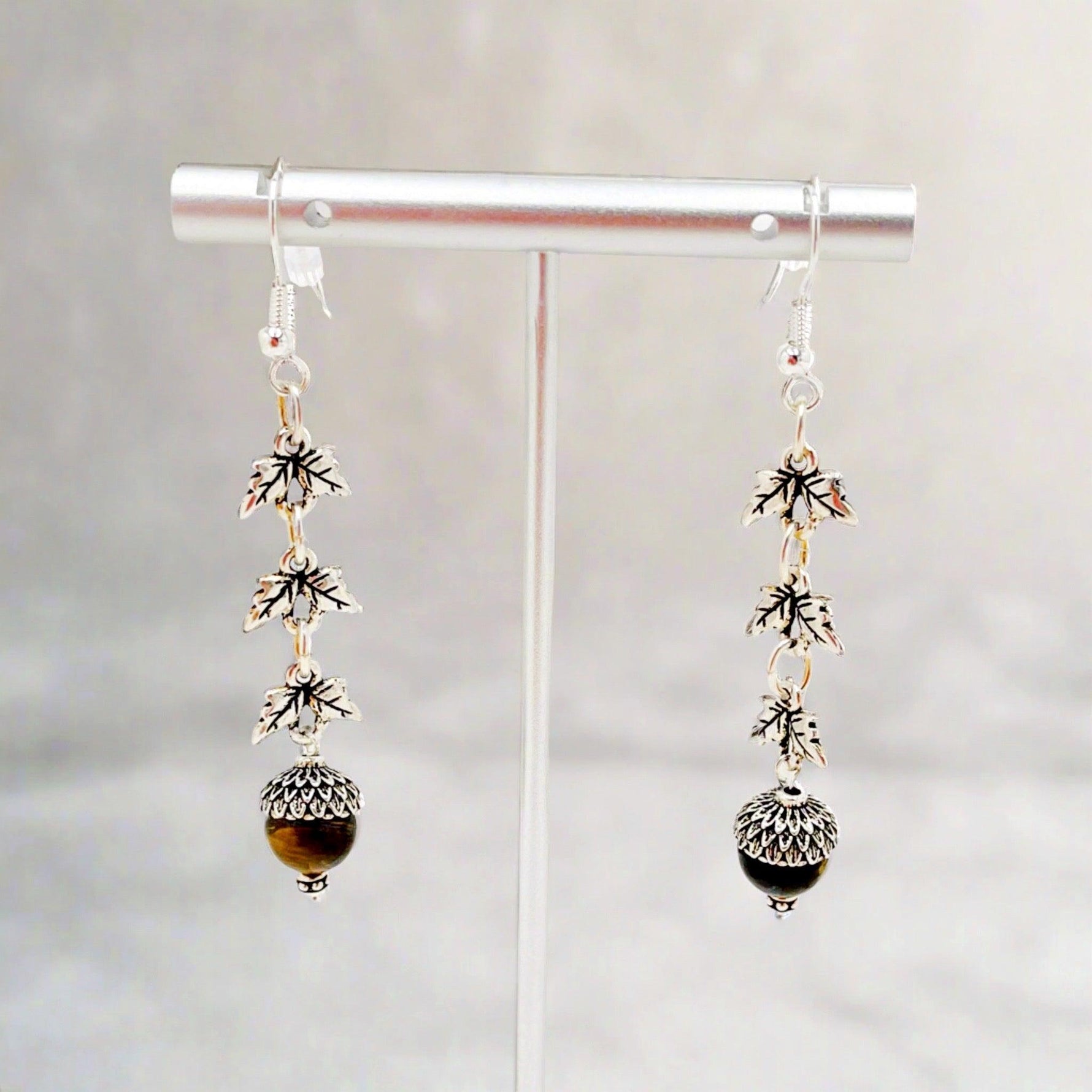 Sterling silver Tiger’s Eye drop earrings displayed on a minimalist stand, highlighting woodland-inspired leaf links and polished gemstone drops with refined artisanal detail.
