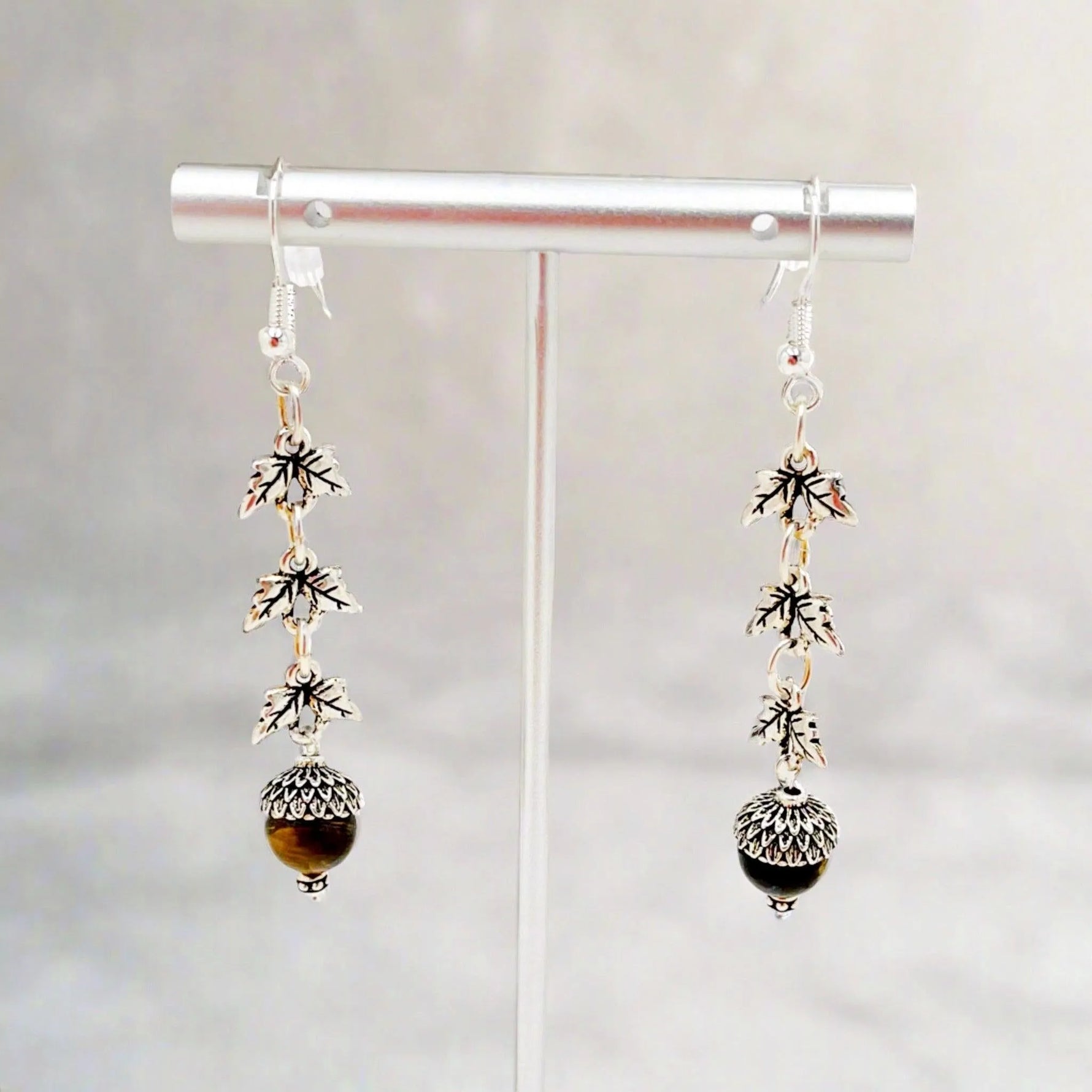 Sterling silver Tiger’s Eye drop earrings displayed on a minimalist stand, highlighting woodland-inspired leaf links and polished gemstone drops with refined artisanal detail.