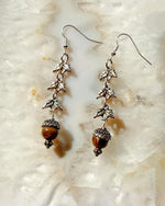 Handcrafted Tiger’s Eye drop earrings in sterling silver with acorn and leaf detailing, photographed on a luminous stone surface showcasing their warm, earthy elegance.