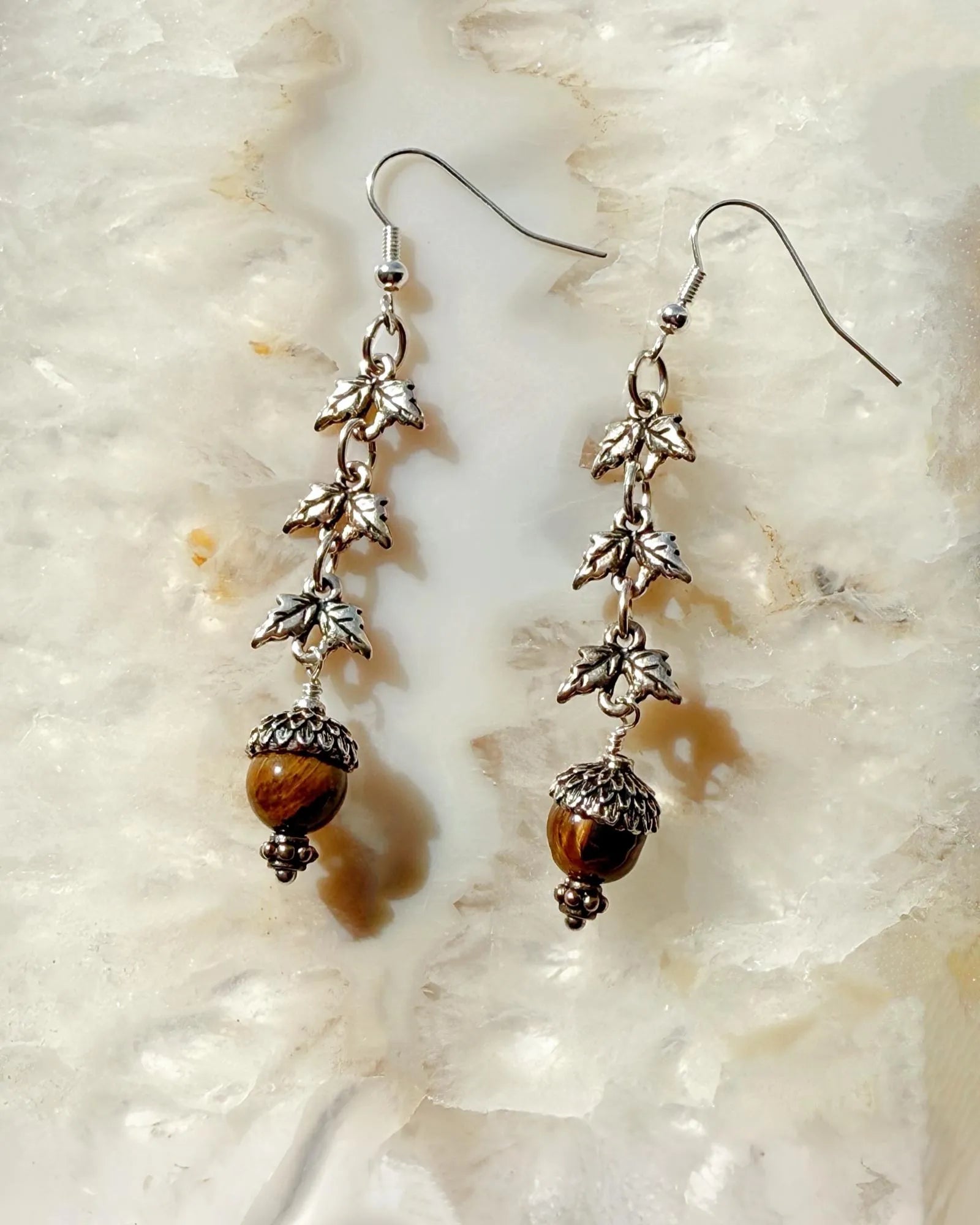 Handcrafted Tiger’s Eye drop earrings in sterling silver with acorn and leaf detailing, photographed on a luminous stone surface showcasing their warm, earthy elegance.