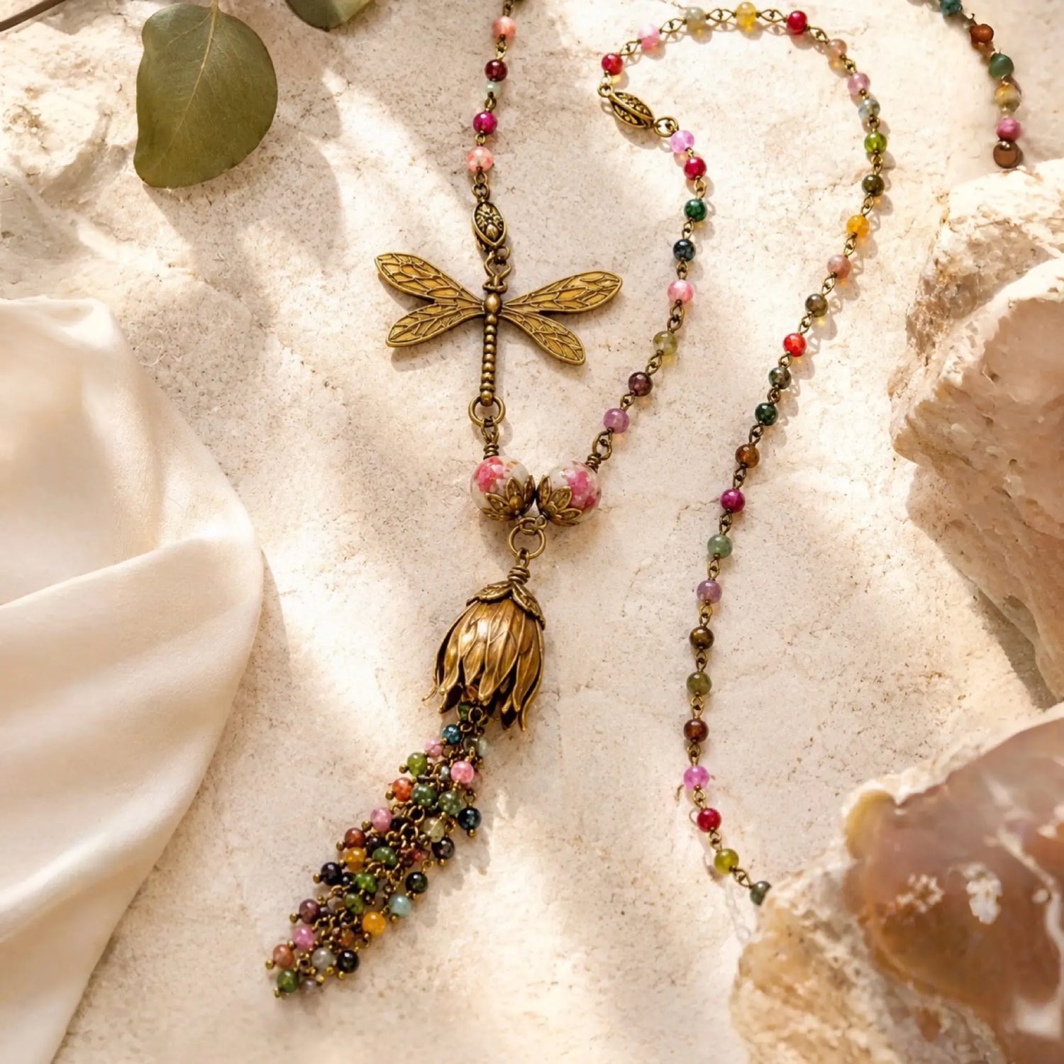 Tulipe necklace featuring watermelon tourmaline gemstones, Vintaj brass tulip focal, dragonfly charm, and gemstone waterfall tassel arranged in a luxury flat lay with warm natural light.