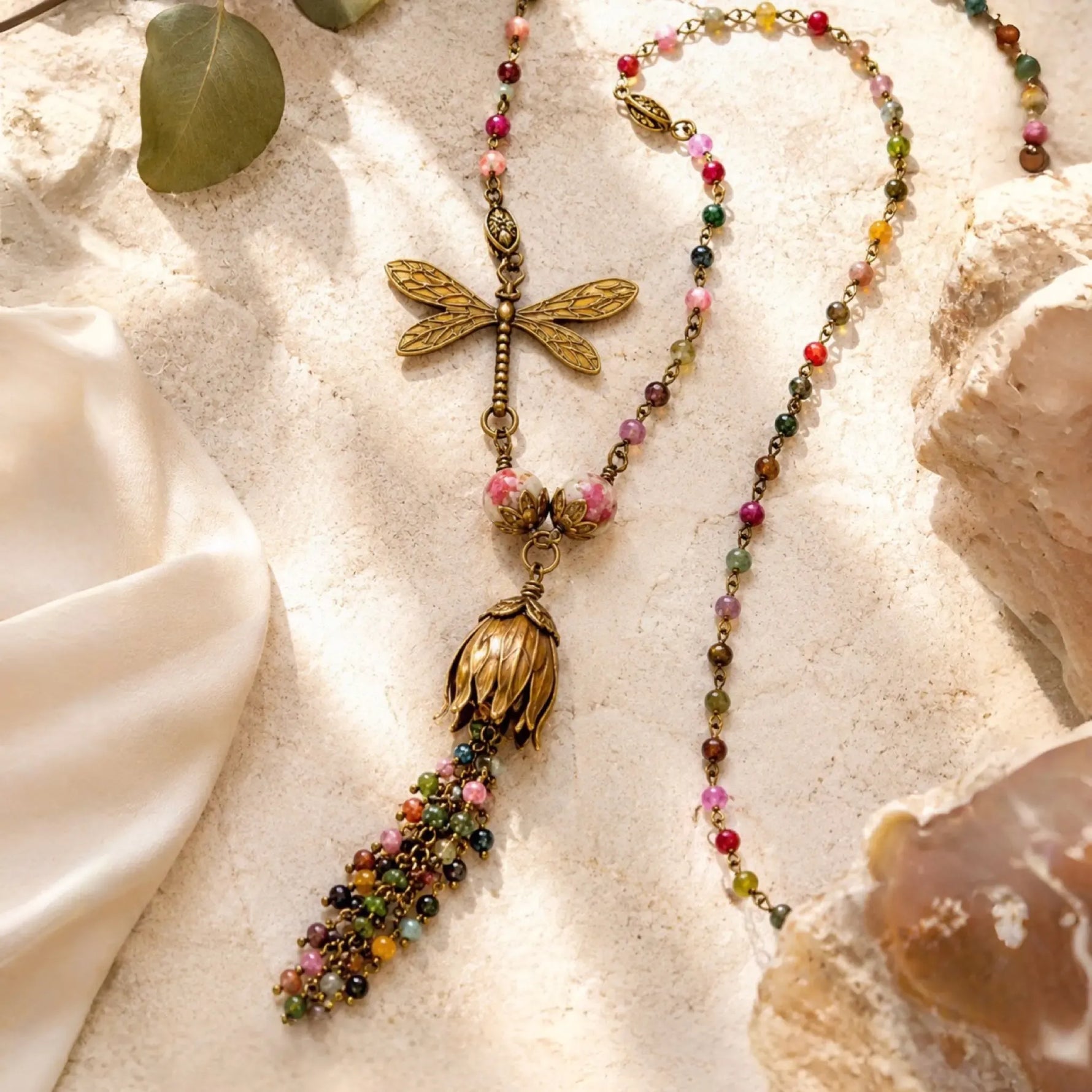Tulipe necklace featuring watermelon tourmaline gemstones, Vintaj brass tulip focal, dragonfly charm, and gemstone waterfall tassel arranged in a luxury flat lay with warm natural light.