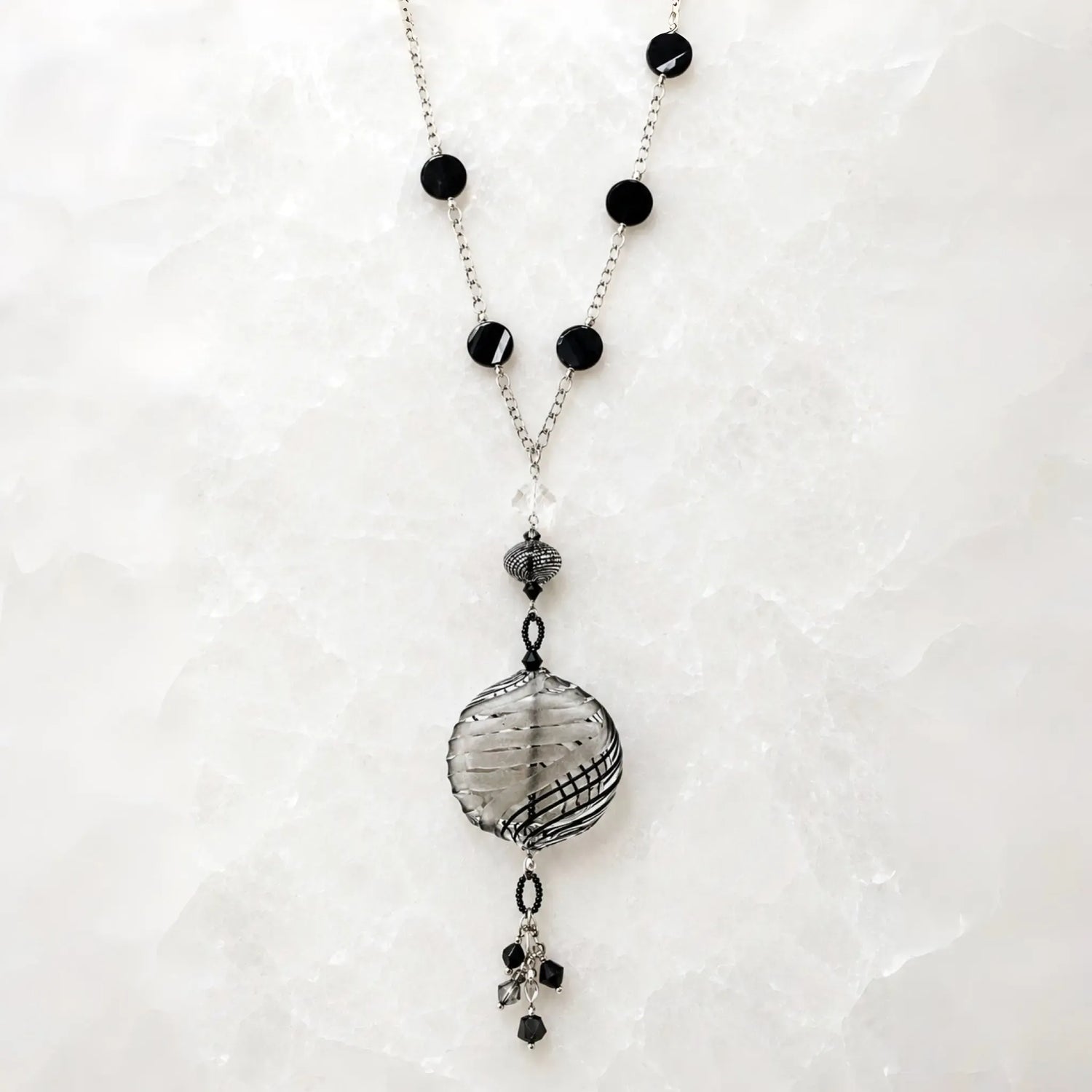 Venezia Murano glass necklace handcrafted in Canada with sterling silver chain, black Murano glass stations, artisan Murano pendant and crystal tassel on white quartz background.